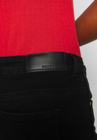 Jeans de mezclilla negros con una textura suave, con un parche de cuero negro en la cintura etiquetado como "NOISY MAY". Parte de una blusa de canalé roja es visible.