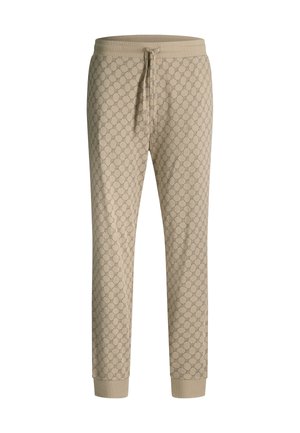 Beige broek met een geometrisch circulair patroon, elastische tailleband, gedragen met een ingestopte witte blouse, waarbij de rechterhand van een persoon naast het lichaam te zien is.