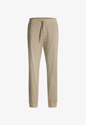 Beige broek met een geometrisch circulair patroon, elastische tailleband, gedragen met een ingestopte witte blouse, waarbij de rechterhand van een persoon naast het lichaam te zien is.