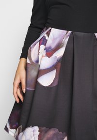 Femme portant un haut noir à manches longues et une jupe foncée avec un grand imprimé floral violet et blanc près de la taille et de l'ourlet.