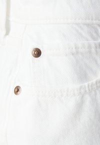 Tissu en denim blanc présentant une texture de côtes visibles, deux poches et des boutons en bronze avec des détails gravés.