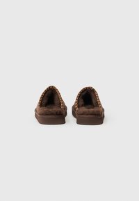 Pantofole marroni con retro aperto, dotate di un morbido interno peloso e un accento decorativo intrecciato lungo il bordo superiore. Sole piatta.