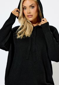 Felpa nera con cappuccio, caratterizzata da una trama a coste, cordino frontale e vestibilità oversize. Maniche lunghe con polsini a coste. Design semplice e moderno.