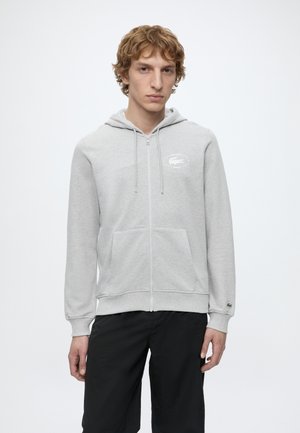 Jonge man met krullend haar die een lichtgrijze zip-up hoodie met voorzakken en een zwarte broek draagt, staand tegen een effen achtergrond.