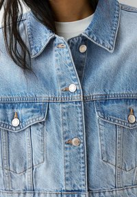 Hellblaue Jeansjacke mit Kragen, silbernen Druckknöpfen, zwei Brusttaschen und sichtbaren Nahtdetails.