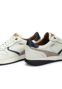 Zapatillas de cuero blanco con acentos en azul marino y beige, paneles de malla, cordones redondos y una suela de goma texturizada negra. Presenta una marca en el costado.