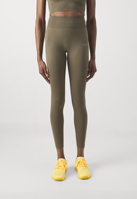 Leggings vert olive avec une taille haute, texture lisse et un détail de logo sur la cuisse gauche, associés à des chaussures de sport jaune vif.
