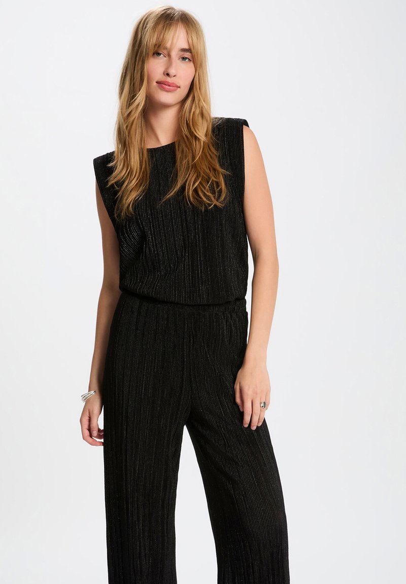 Zwarte, mouwloze jumpsuit met een verticale ribbelstructuur. Elastische tailleband voor comfort. Model staat voor een neutrale achtergrond en toont de outfit.
