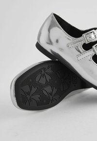 Silberne Lacklederschuhe mit Schnallen, die eine strukturierte schwarze Außensohle mit floralem Muster aufweisen. Abgerundete Form und glatte Oberfläche.