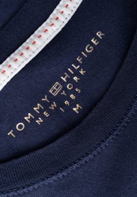 Marinblått tyg med en slät yta, med en vit krage med röd sömnad. Etiketten säger "TOMMY HILFIGER NEW YORK 1985" i guld. Storlek M.