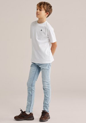 Jongen staand met handen achter de rug, draagt een wit T-shirt, lichtblauwe spijkerbroek en bruine sneakers, tegen een effen achtergrond.