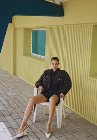 Black leather oversized jacket, patterned blouse, and black shorts. White slingback heels, seated on a white chair against a yellow wall.