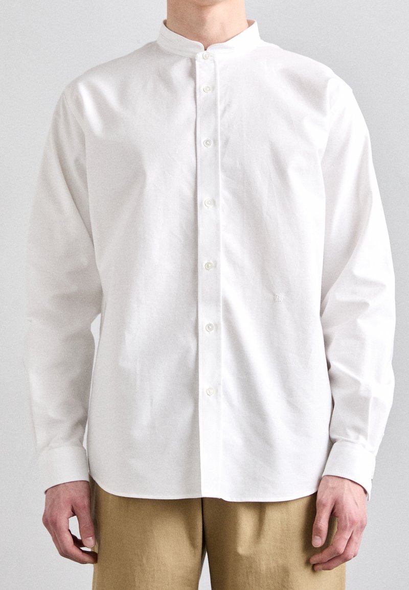 Homme portant une chemise blanche unie à boutons avec col mandarin et un pantalon beige, se tenant devant un fond uni.