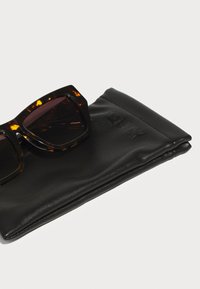 Occhiali da sole tartarugati con montatura angolare e lenti scure, appoggiati su un astuccio in faux pelle nera impresso con la scritta "QUAY."
