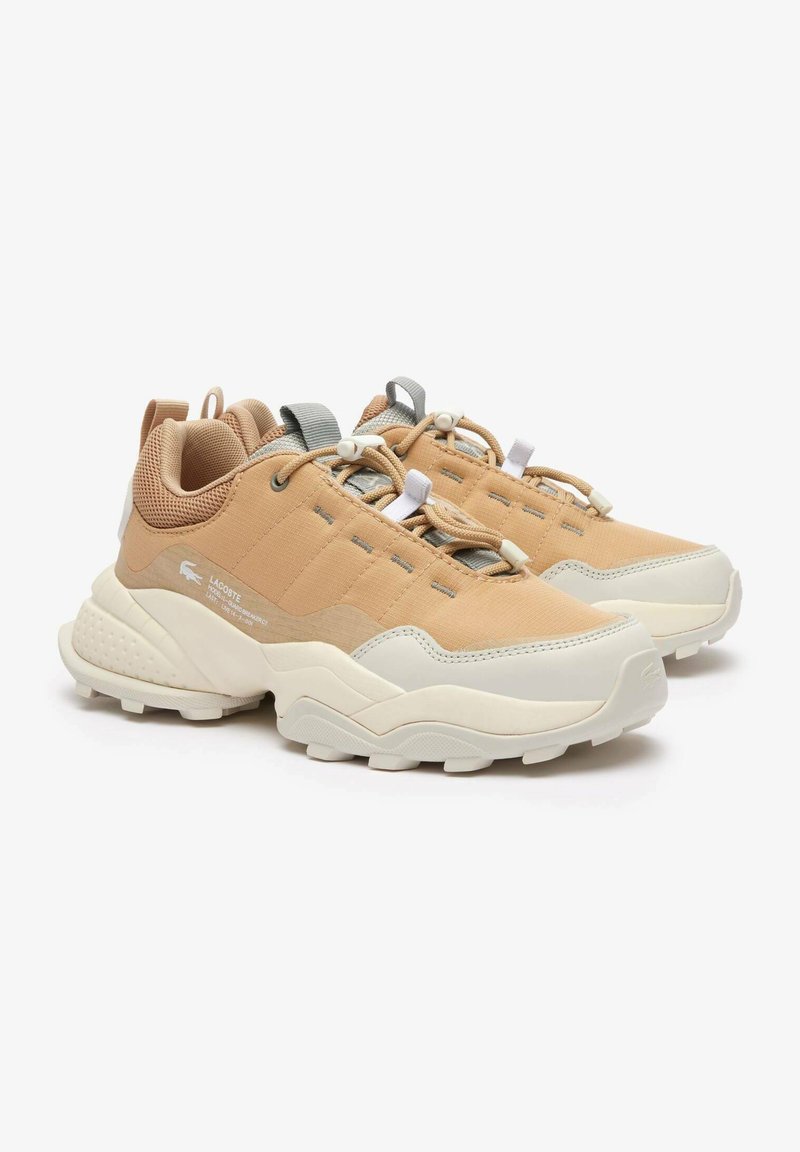 Beżowe i kremowe niskie sneakersy Lacoste z grubą podeszwą, uchwytami, elastycznymi sznurówkami oraz subtelnym logo marki na boku i palcach.