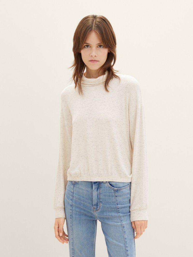 Maglietta a manica lunga color beige chiaro con collo alto, caratterizzata da un fit rilassato e una trama leggermente screziata, abbinata a jeans blu chiaro.