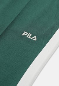 Grønt stof med hvid sideliste og broderet FILA-logo i hvid tråd på tekstureret materiale.