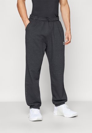 Mand iført mørkegrå joggingbukser med elastiske manchetter og hvide sneakers, stående med den ene hånd i lommen mod ensfarvet baggrund.