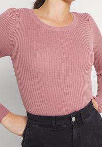 Pull rose côtelé à manches longues avec un col rond. Caractérisé par des manches légèrement bouffantes et associé à un pantalon noir taille haute.