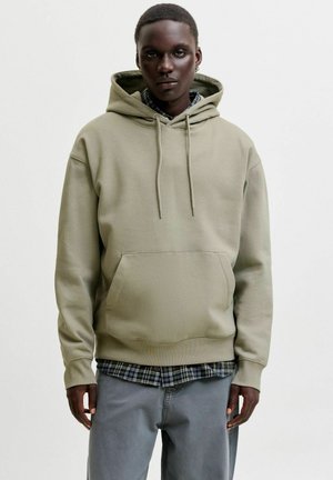 Junge Person trägt beige Kapuzenpullover über kariertem Hemd und grauer Hose, steht vor hellem, einfarbigem Hintergrund.