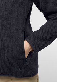 Giacca in fleece nera con tessuto testurizzato, una tasca frontale con zip e un orlo curvo. Presenta un design dei polsini attillato e un branding sottile.