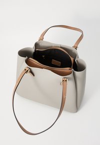 Sac cabas gris clair avec poignées en cuir fauve, ouverture supérieure montrant des compartiments intérieurs zippés noirs et fauves sur fond blanc.