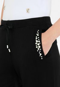 Sorte bukser med en dekorativ lomme, der har perler i varierende størrelser i hvid. Stoffet ser blødt ud; taljebåndet har en snor.