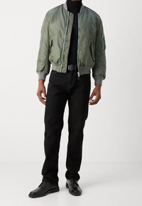 Blouson aviateur vert olive avec une fermeture éclair, des poignets côtelés et une poche latérale. Porté sur un col roulé noir et associé à un jean noir et des chaussures noires.