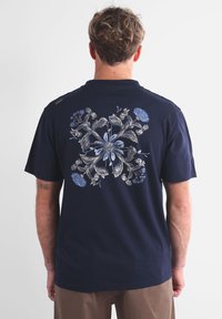 Homme avec des cheveux courts et bouclés, tourné de dos, portant un t-shirt bleu marine avec un design floral symétrique en beige et bleu clair dans le dos.