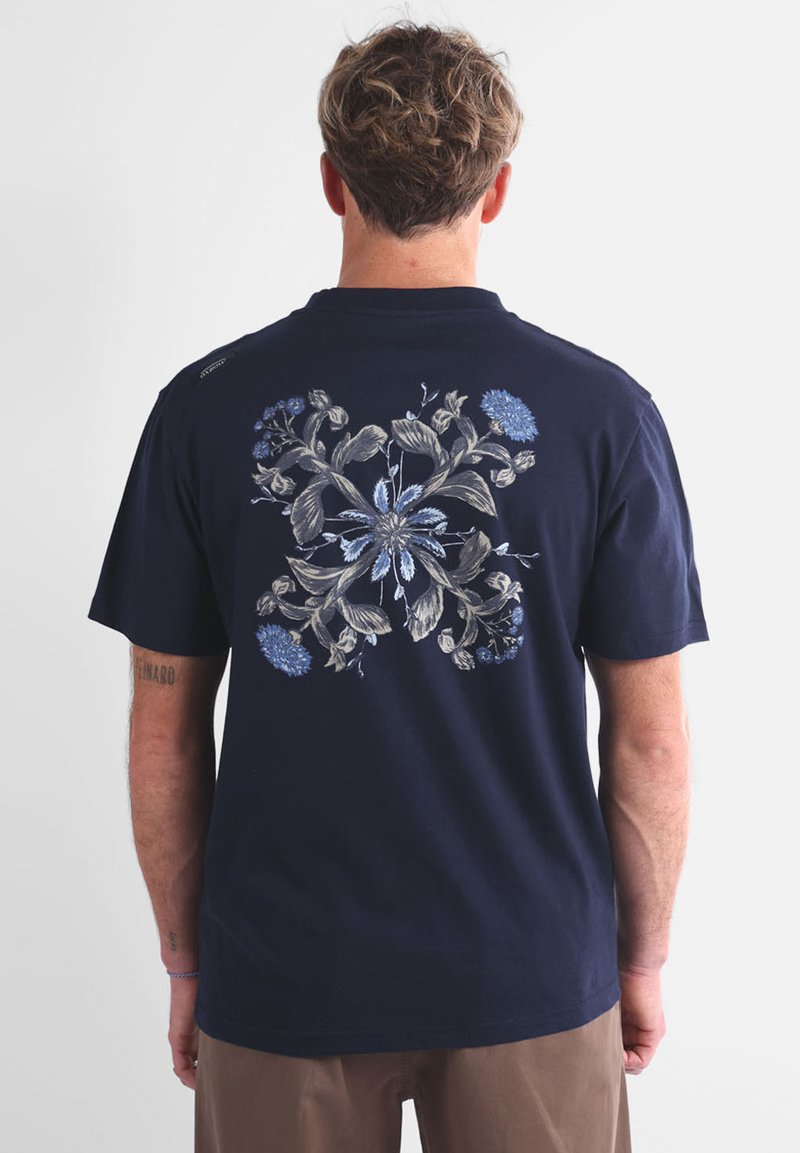 Homme avec des cheveux courts et bouclés, tourné de dos, portant un t-shirt bleu marine avec un design floral symétrique en beige et bleu clair dans le dos.