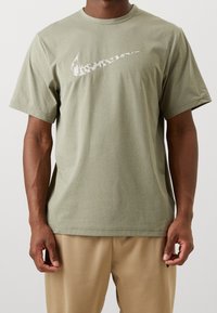 Oliwkowa bawełniana koszulka z krótkim rękawem, z dużym, fakturowanym białym logo Nike na klatce piersiowej. Okrągły dekolt, luźny krój.