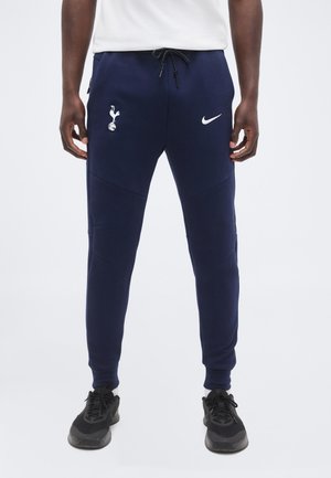 Άνδρας που φοράει σκούρο μπλε φόρμα Nike με το λογότυπο της Tottenham Hotspur, μαύρα αθλητικά παπούτσια και λευκό πουκάμισο σε απλό φόντο.