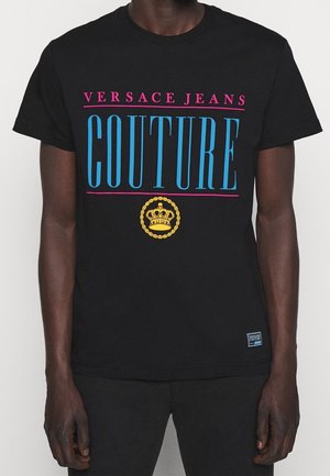 T-shirt noir avec inscription rose "Versace Jeans", grand "Couture" bleu et emblème de couronne jaune porté par une personne à la peau foncée.