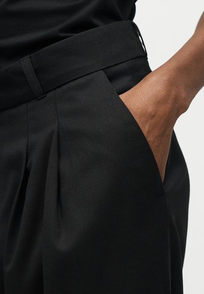 Χέρι στην τσέπη μαύρου ψηλόμεσου παντελονιού με πιέτες που φοριέται με μαύρο τοπ μέσα στη μέση σε καφέ δέρμα.