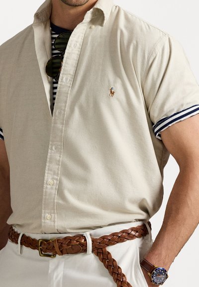 Short-sleeve beige cotton shirt with a light texture, featuring a small embroidered logo and striped navy accents on the sleeves.