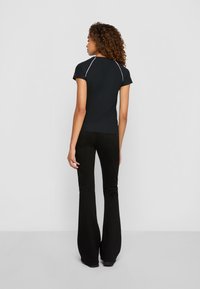 Vrouw met krullend haar, gekleed in een zwart T-shirt met korte mouwen en zwarte uitlopende broek, staande met de rug naar de camera tegen een effen grijze achtergrond.