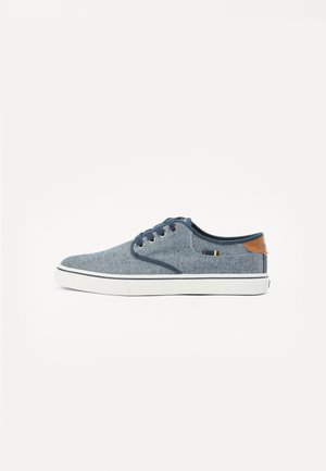 Blå denim sneakers med en glat tekstur, hvid gummisål og brunt læderdetalje på hælen. Har fire huller til snørebånd.