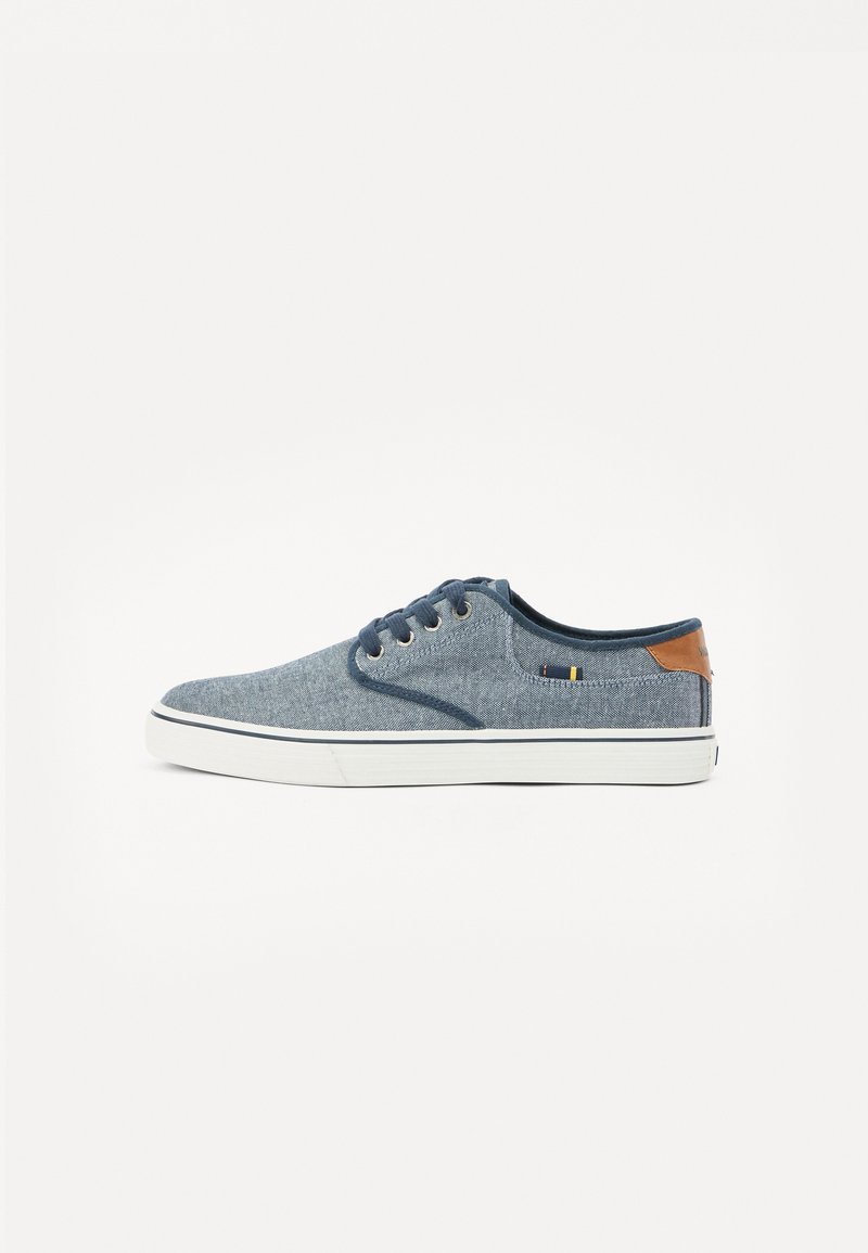 Sapatilhas de denim azul com uma textura suave, sola de borracha branca e detalhe de couro castanho no calcanhar. Apresentam quatro ilhós para os atacadores.