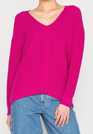 Personne portant un pull en maille rose vif à col en V et un jean taille haute bleu clair, debout avec une main reposant sur la cuisse.