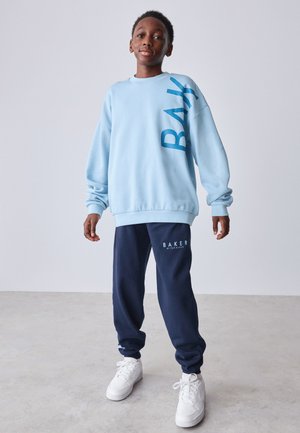 Poiss seisab helesinises üleulatuvas dressipluusis, millel on kiri "BAY", tumesinistes dressipükstes kirjaga "BAKER" ja valgetes tossudes hallil põrandal ja seinal.