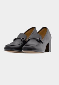 Μαύρα δερμάτινα μοκασίνια με τακούνι, με στρογγυλεμένη μυτερή μύτη, χαμηλό τετράγωνο τακούνι και διακοσμητικό μεταλλικό εξάρτημα στο πάνω μέρος. Ομαλή υφή, μίνιμαλ σχεδιασμός.