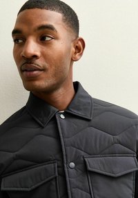 Czarna pikowana kurtka z kołnierzem, zatrzaskami i dwoma kieszeniami na piersi z geometrycznym wzorem. Gładka faktura i nowoczesny design.