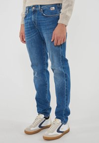 Persona che indossa jeans in denim blu e sneaker bianche con dettagli blu navy e beige, in piedi contro uno sfondo chiaro e semplice.
