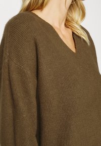 Brun V-ringad tröja med texturerad stickning, som har löst sittande ärmar och en mjuk, varm känsla. Sidasyn som framhäver passform och tyg.