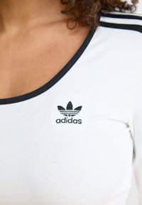 White fitted shirt with a black neckline and three thin black stripes on the shoulders. Features a black Adidas logo on the chest.