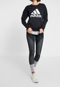 Felpa nera con grande logo bianco Adidas; abbinata a leggings neri e grigi a fantasia. Le scarpe sono sneakers da corsa grigie chiare.