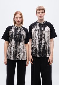 Μαύρα και λευκά γραφικά t-shirts με ραβδωτό μοτίβο, που διαθέτουν μανίκια ράγκλαν. Και οι δύο φορούν άνετο, φαρδύ μαύρο παντελόνι.