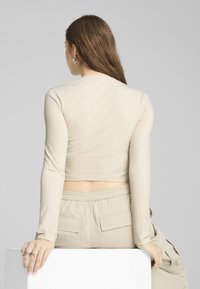 Beige pitkähihainen crop-top, joka on valmistettu sileästä kankaasta. Tässä on pyöreä pääntie ja istuva malli, yhdistettynä vaaleanbeigeihin cargo-housuihin.
