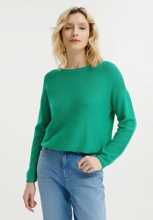 Grüner Strickpullover mit weitem Ausschnitt und langen Ärmeln, kombiniert mit hellblauen Jeans. Der Stoff hat eine gerippte Textur.