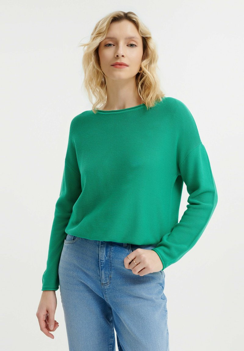 Grüner Strickpullover mit weitem Ausschnitt und langen Ärmeln, kombiniert mit hellblauen Jeans. Der Stoff hat eine gerippte Textur.